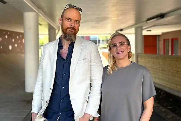 En man med vit kavaj, solglasögon på huvudet och skägg står bredvid en ljushårig kvinna i en tunnel utomhus.